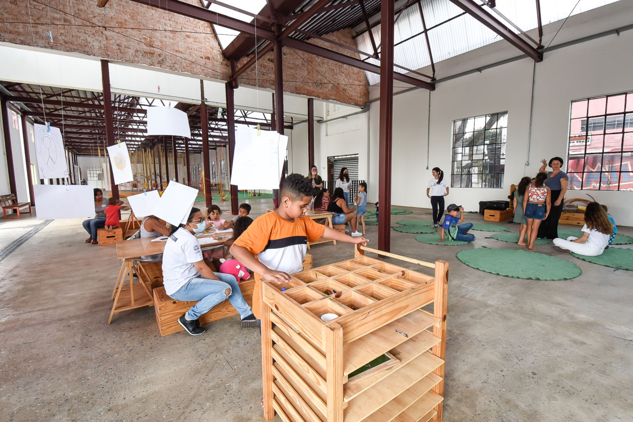 Crianças e adultos realizando atividades de pintura e outras brincadeiras na Fábrica das Infâncias Japy, onde se veem bancadas e mesas de madeira, tapete de grama sintética no chão e folhas com desenhos penduradas do teto