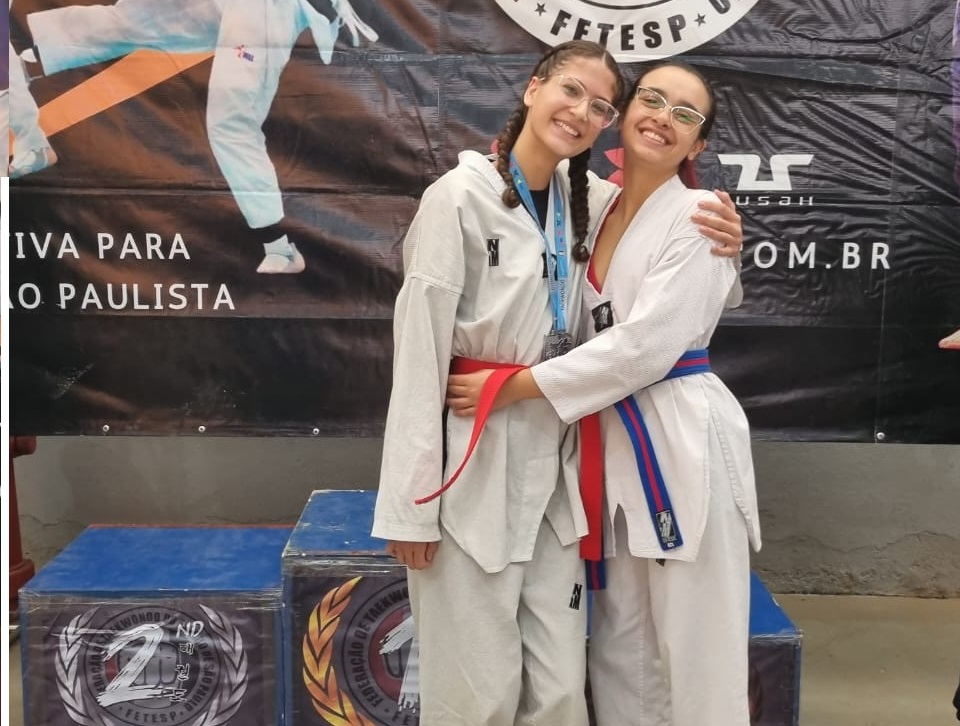 Foto mostra duas meninas atletas do Taekwondo usando kimono branco e faixa verde, se abraçando, com medalhas do peito. 