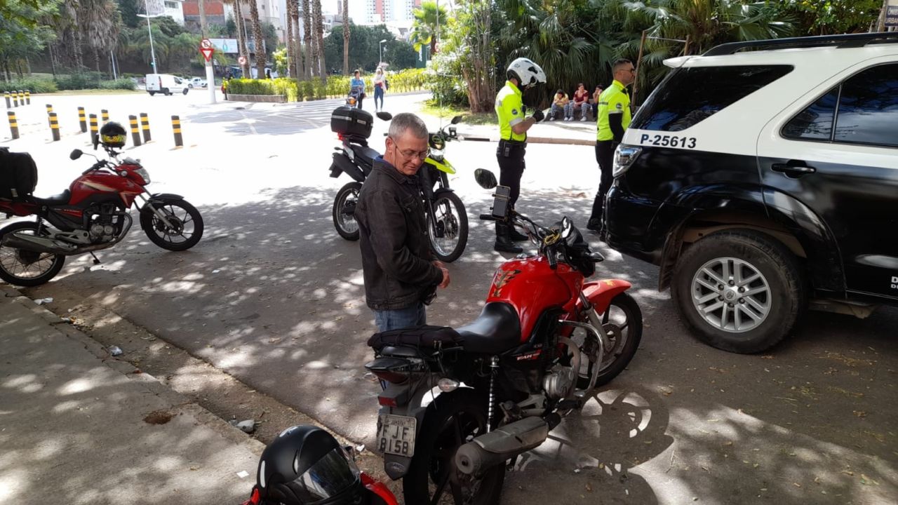 Motociclista é parado para fiscalização de documentos e cadastro nos aplicativos de entrega. Ele está em pé ao lado de sua motocicleta vermelha