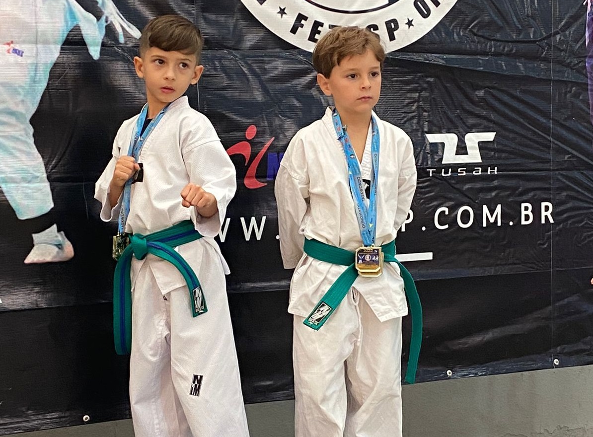 Foto mostra dois meninos atletas do Taekwondo usando kimono branco e faixa verde, com medalhas do peito. 