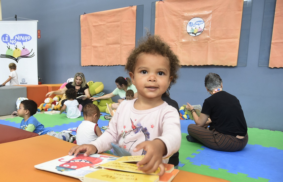 Criança em pé apoiada em bancada colorida, folheando livro com ilustrações, usando blusa com de rosa com desenho de unicórnio, com mulheres e crianças sentadas ao fundo, brincando sobre piso emborrachado