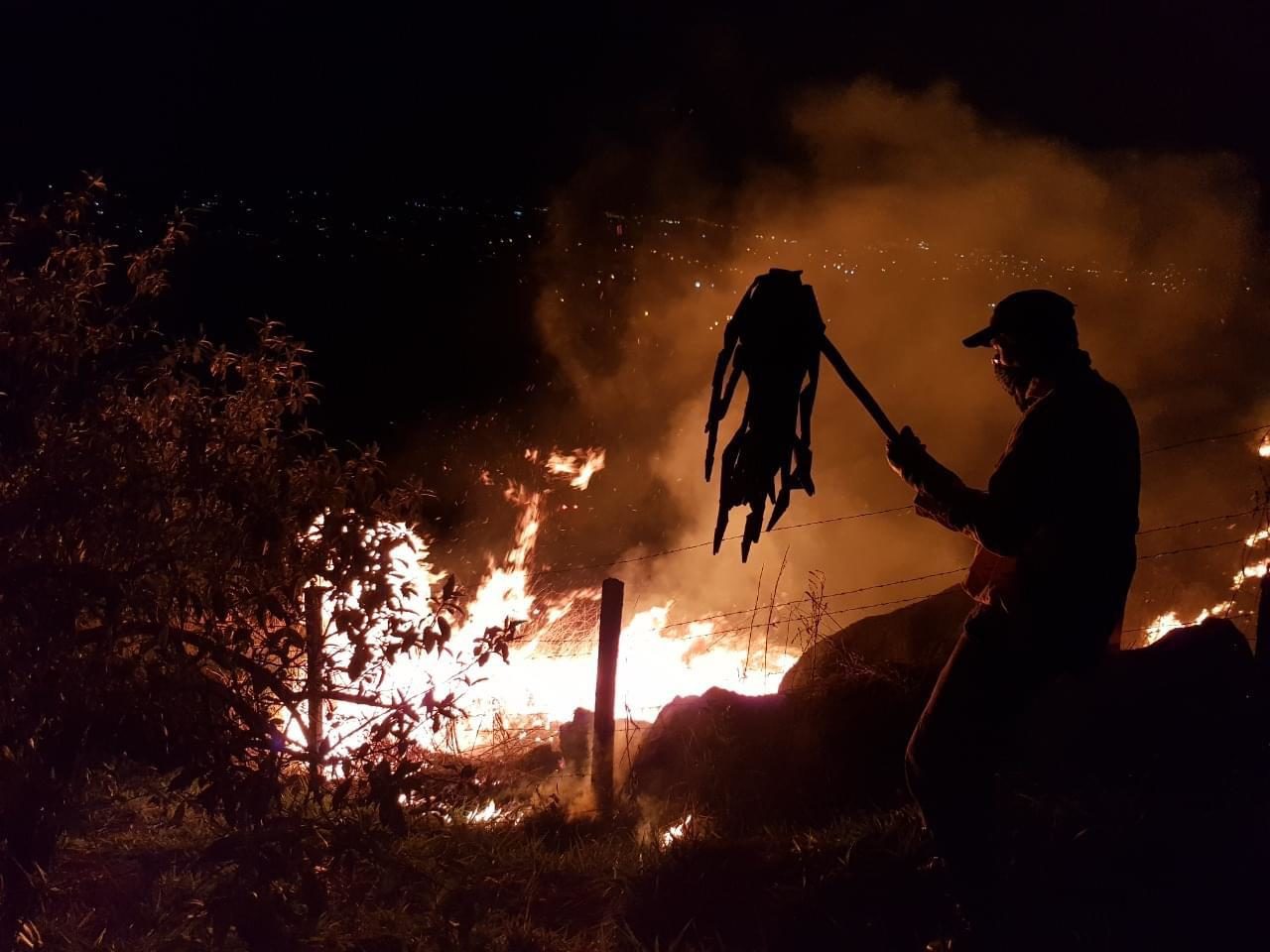 DESCRIÇÃO DE IMAGEM: foto noturna de incêndio. Labaredas são combatidas por homem, com equipamento abafador