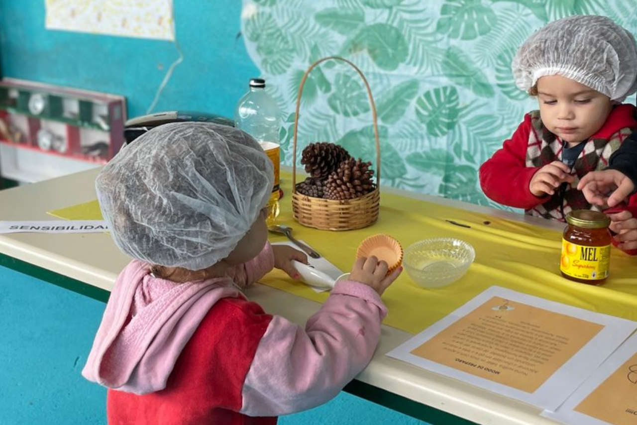 Com um fundo de papel de parede de plantas e parede azul, no foco, são duas meninas pequenas de touca branca, com as mãos na mesa, onde estão os ingredientes para a receita de cupcake.