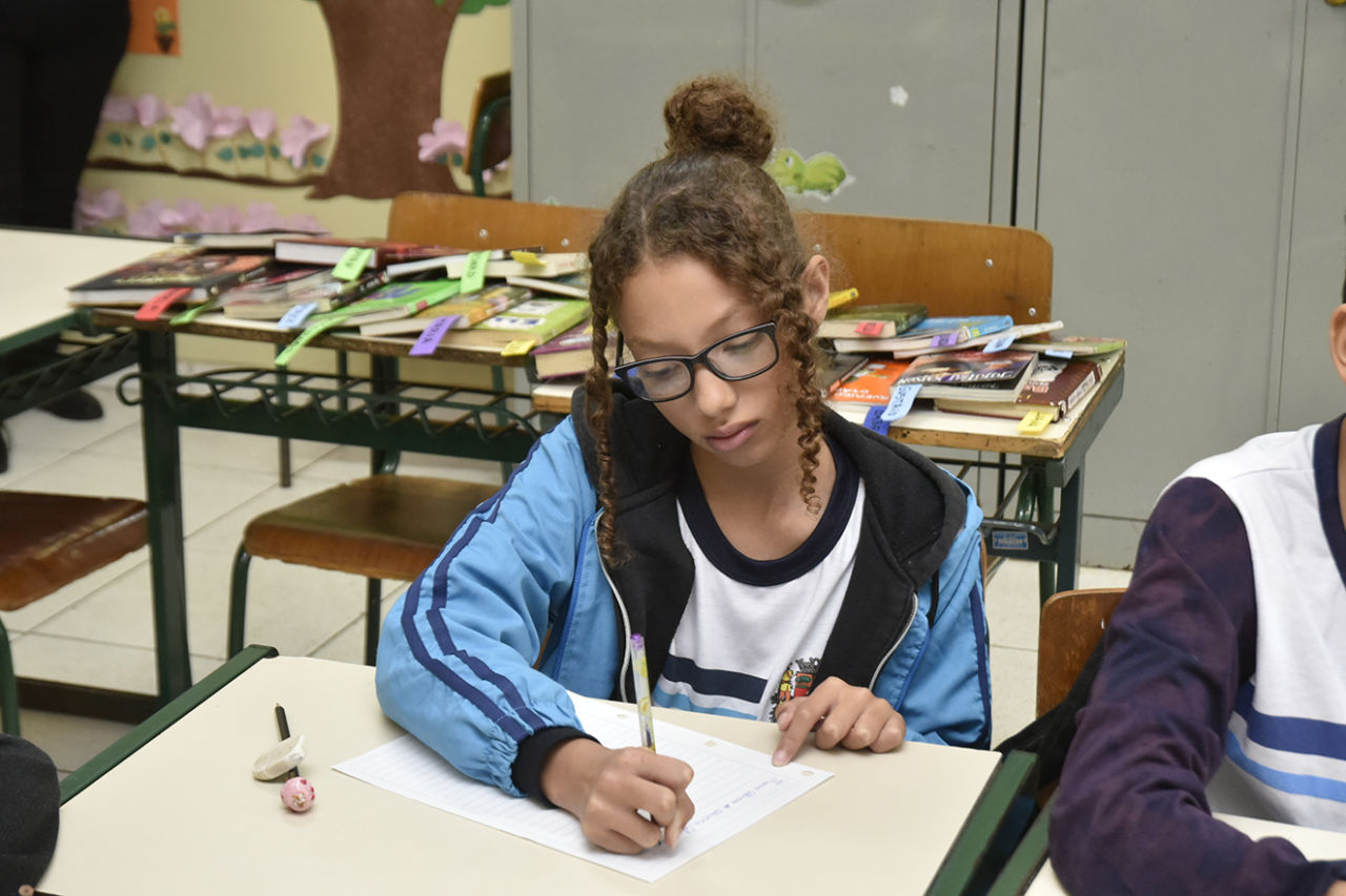 Uma menina, de coque e franja solta, óculos preto e jaqueta do uniforme, escreve em uma folha branca, apoiada em uma mesa, com lápis e borracha ao lado. No fundo, encontram-se duas mesas cheias de livros, dentro da sala de aula.