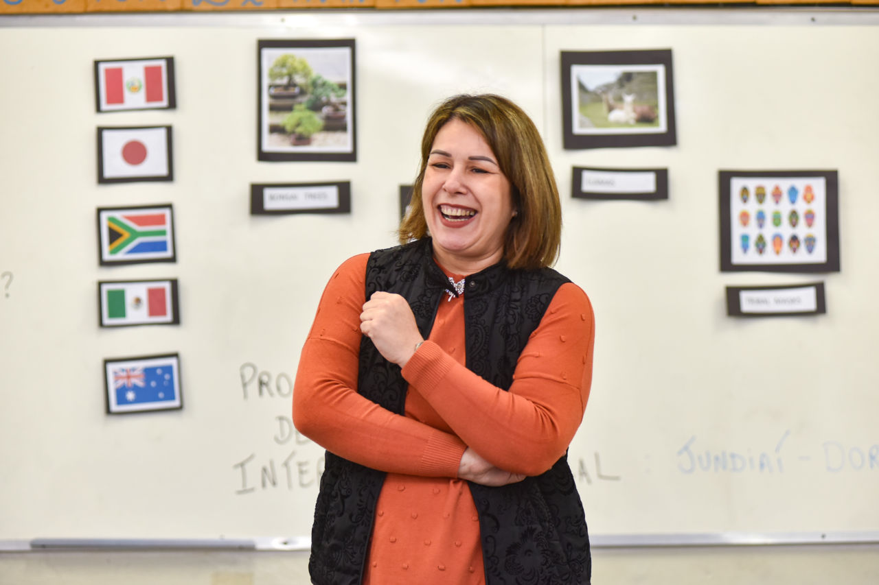 A foto é da professora, onde aparece com uma blusa laranja e colete preto, sorrindo, com a lousa de fundo.