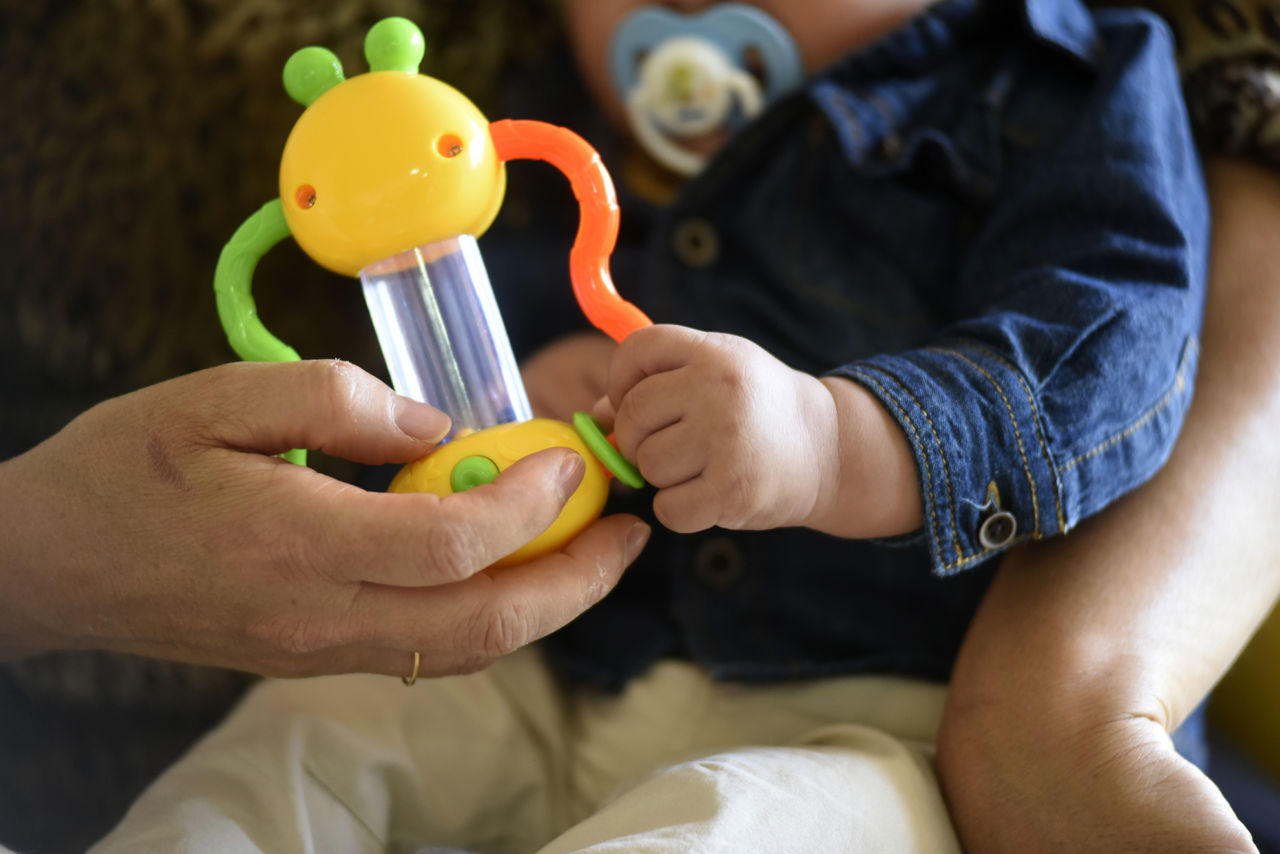 Detalhe de bebê de colo, com roupas de manga comprida e calça, com chupeta na boca, segura brinquedo infantil colorido de chocalho, sobre colo de pessoa adulta, segurando o brinquedo com aliança na mão.