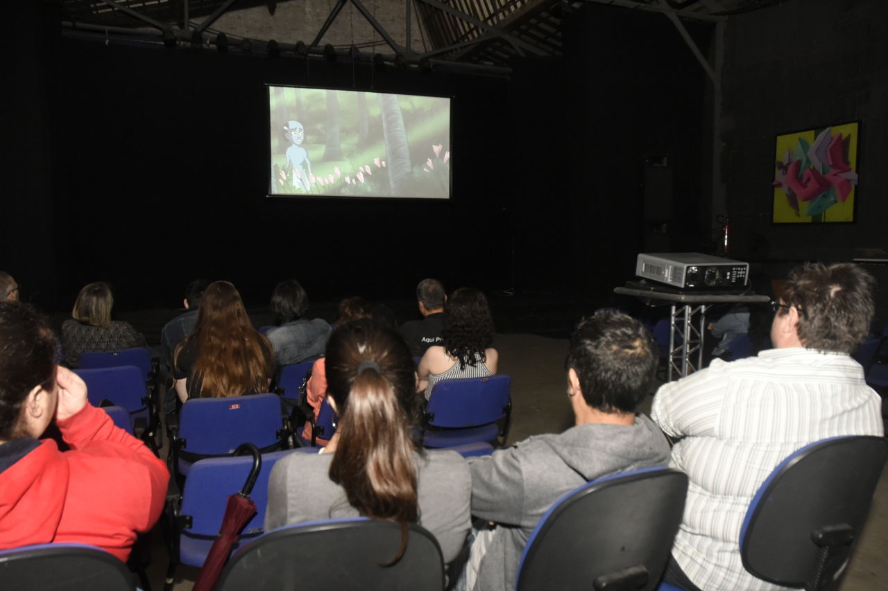 Sala Jundiaí do Complexo Fepasa com pessoas de costas, sentadas sobre cadeiras estofadas azuis, assistindo a um vídeo projetado em telão