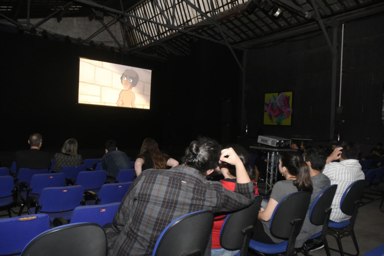 Sala Jundiaí do Complexo Fepasa, com pessoas sentadas sobre cadeiras de estofamento azul, assistindo a um desenho projetado sobre telão disposto sobre o palco
