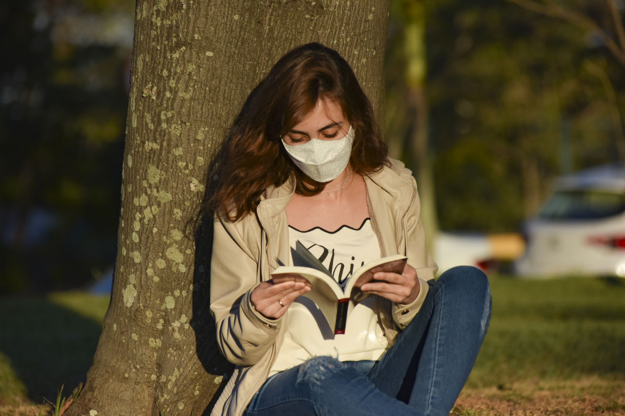 Mulher de cabelos longos, castanhos e lisos, sentada sobre gramado e apoiada em tronco de árvore, lendo um livro, com uma máscara no rosto