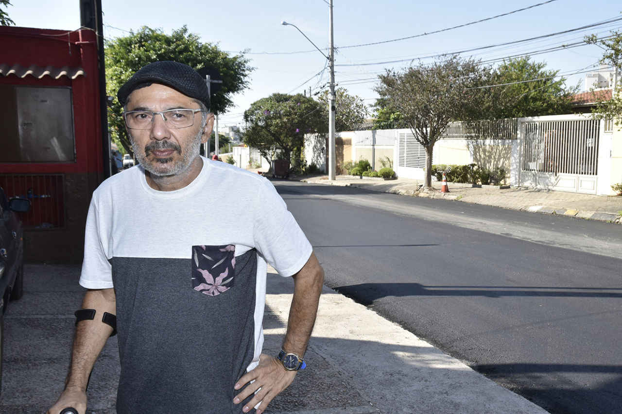 O comerciante Sebastião está na frente da avenida asfaltada, com casas ao fundo; ele veste uma camisa branca e preta, usa óculos e boina preta