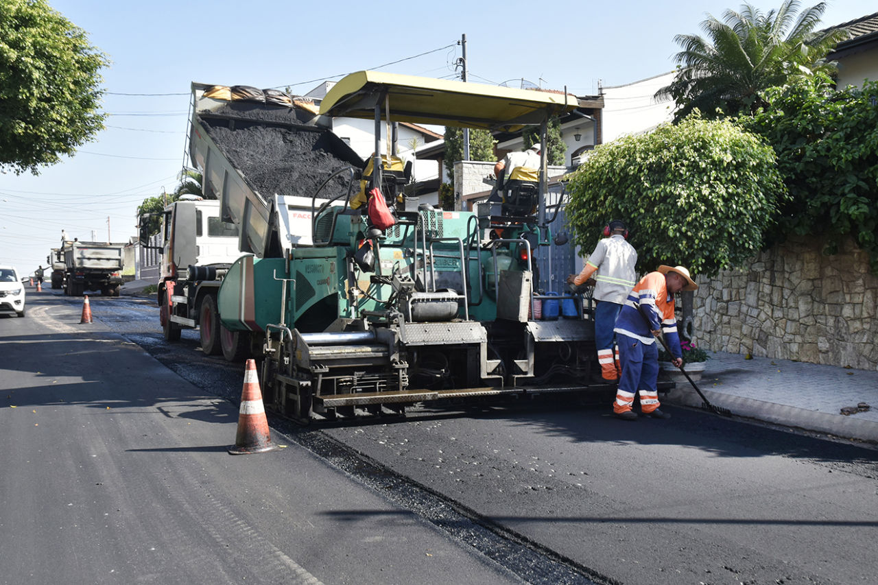 A imagem mostra uma máquina despejando a massa asfáltica na avenida enquanto homens espalham o material no chão; à frente, um caminhão despeja o asfalto na mesma máquina