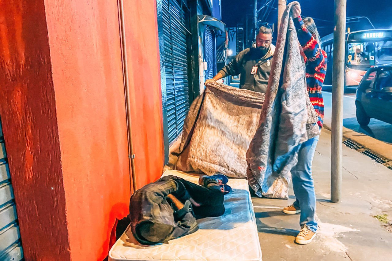 Um homem e uma mulher em pé estendem cobertores sobre pessoa deitada sobre colchão em calçada de rua, por onde passam carro e ônibus