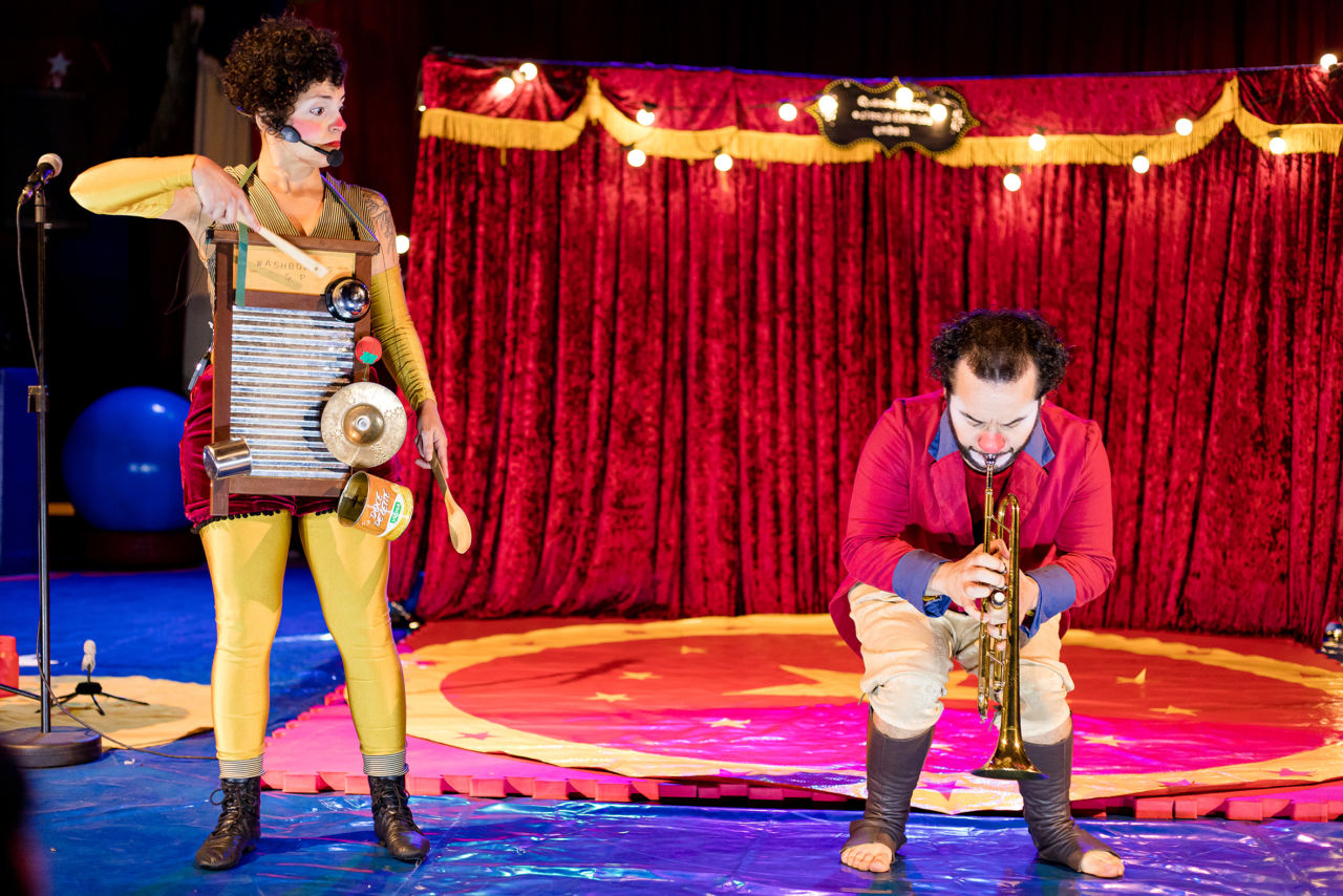 Uma mulher e um homem, vestidos de palhaço e segurando instrumentos musicais, apresentando-se em cenário de circo, com lâmpadas e cortina vermelha e detalhe amarelo ao fundo