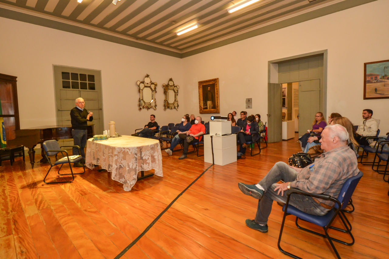 Sala do Museu Solar do Barão, com piso em madeira, recebe visitantes, sentados em cadeiras, para a explanação do gestor Sinésio. 