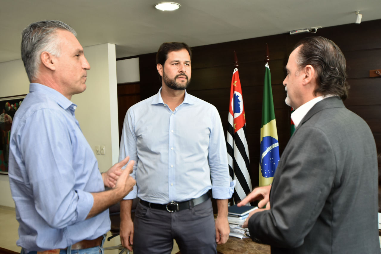 Em pé, com bandeiras ao fundo, no gabinete do prefeito, estão Guilherme Campos, o prefeito Luiz Fernando e o gestor da Casa Civil, Gustavo Maryssael; eles formam uma roda e conversam