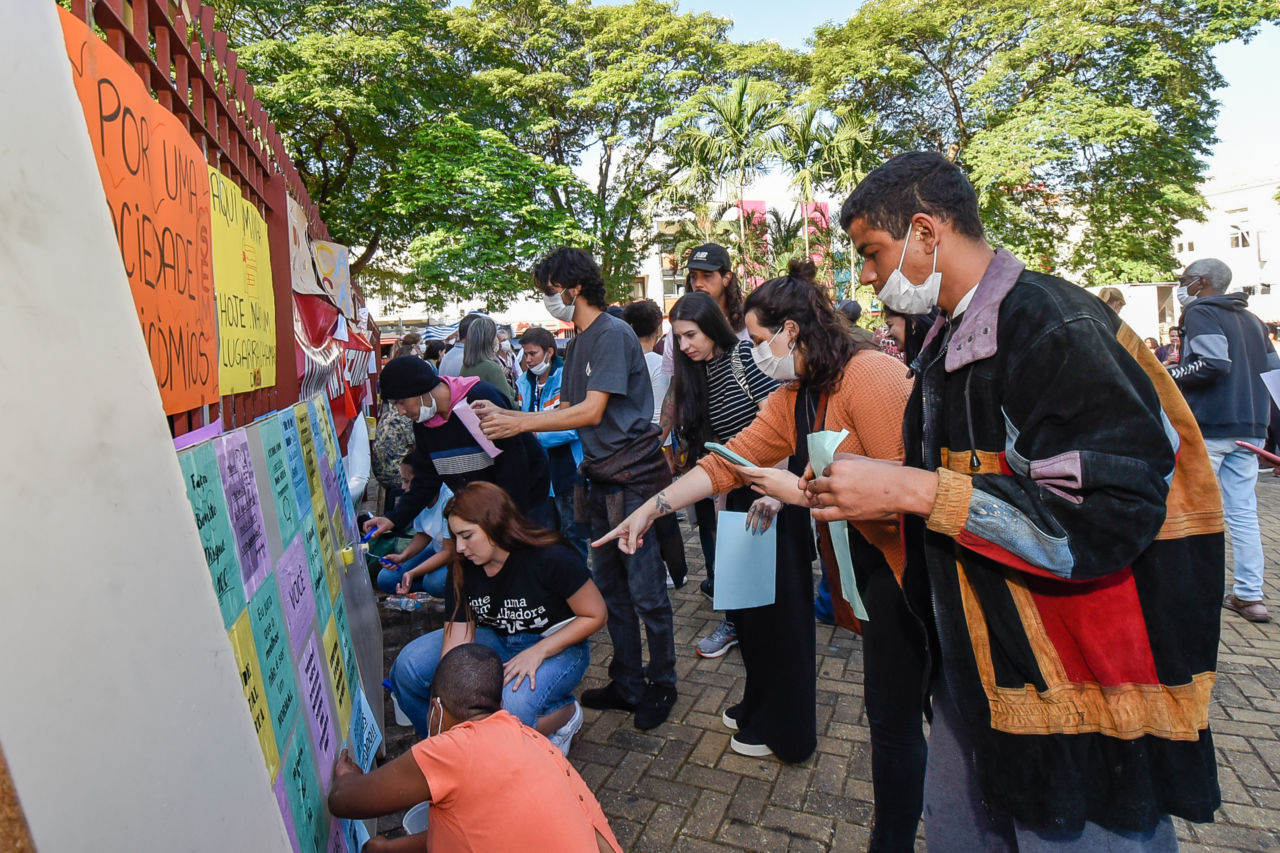 Diversas pessoas em pé e sentadas, em frente a um painel apoiado em grades, onde são afixados cartazes