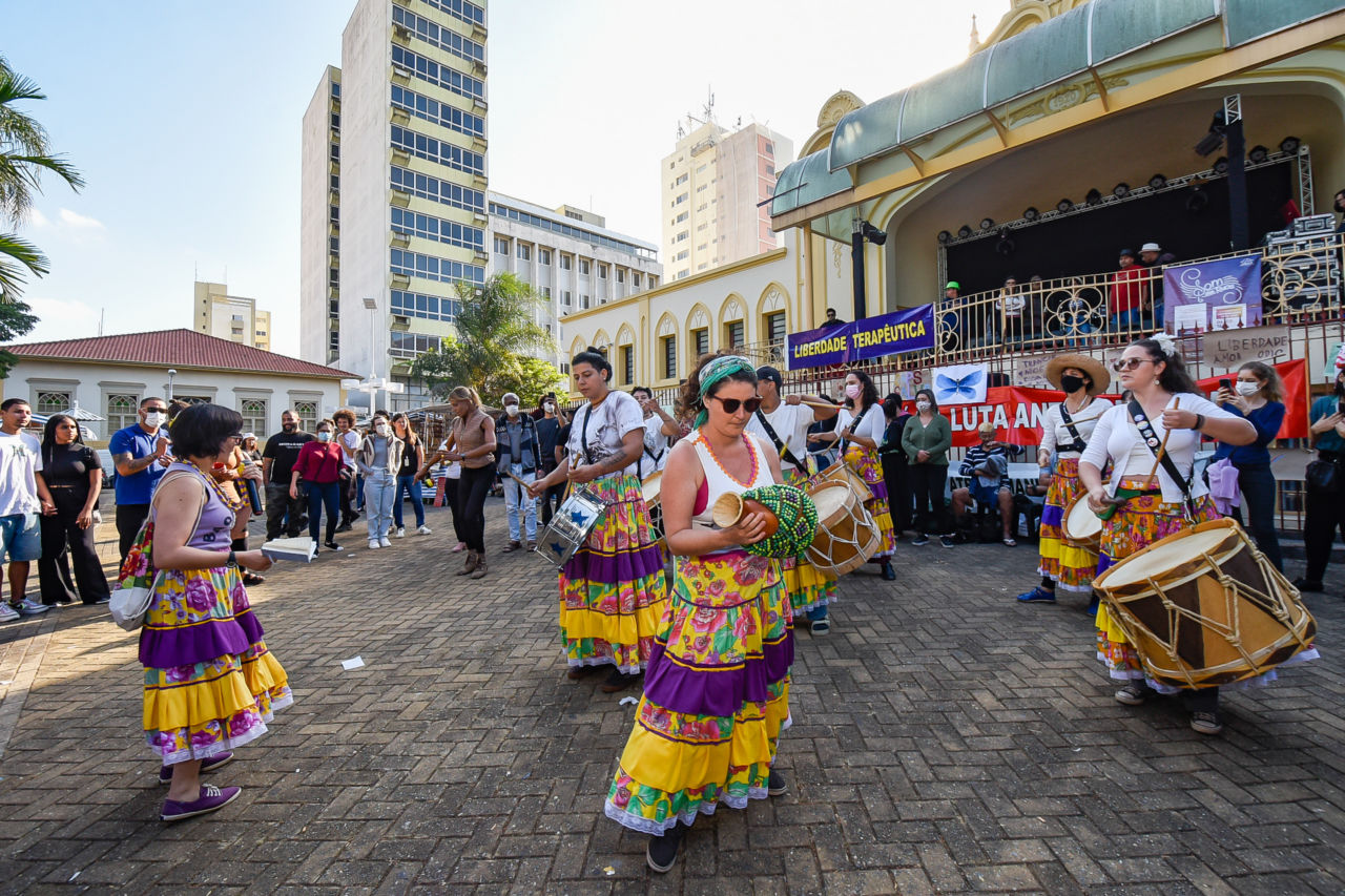 Grupo de pessoas diversas dançando maracatu, com grupo vestido com saias coloridas amarelas, roxas e estampas, segurando instrumentos de percussão, em praça com palco, árvore e prédios ao fundo