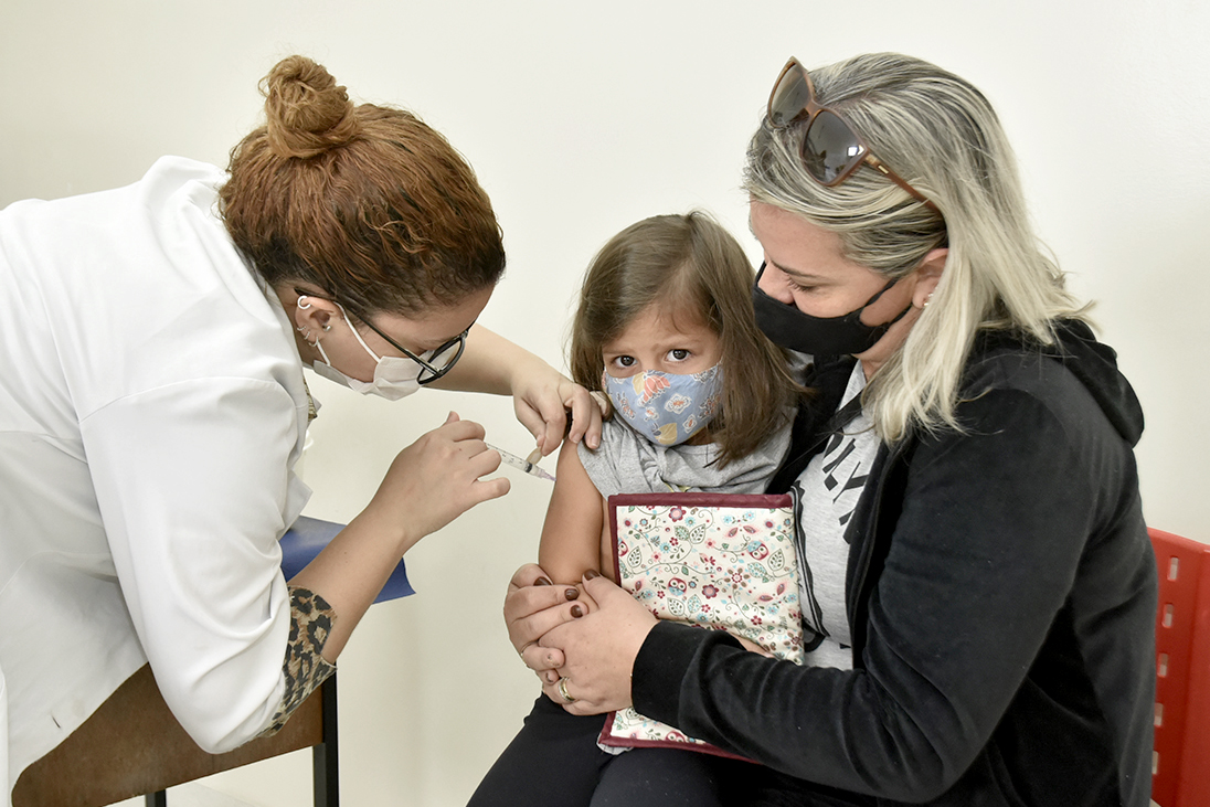isado0ra, de 4 anos, usando máscara, recebe a vacina de uma profissional da saúde. Ela está sentada no colo da mãe e olhando para a câmera