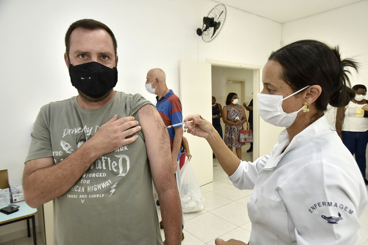 DESCRIÇÃO DA IMAGEM: técnica de enfermagem vestida com jaleco branco e máscara branca realiza a aplicação de vacina em homem, vestido de camiseta verde cinza e máscara preta