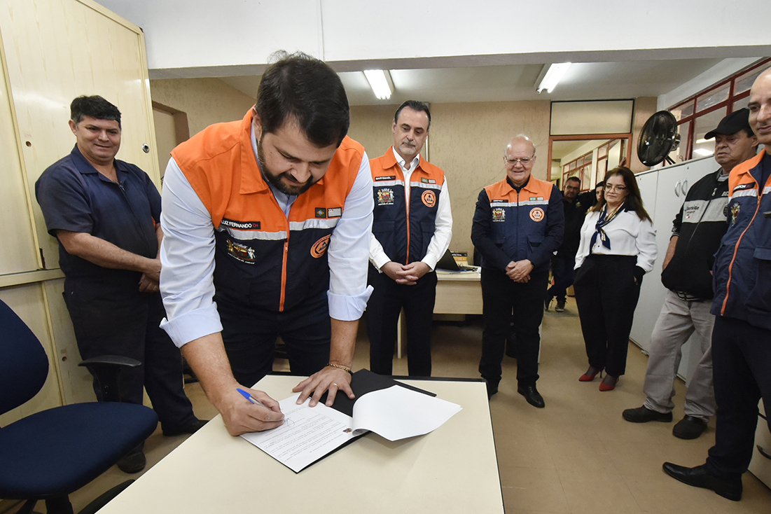 Prefeito Luiz Fernando, vestindo colete azul e laranja de Defesa Civil, assina o decreto que institui a que institui a Medalha da Defesa Civil em Jundiaí.  