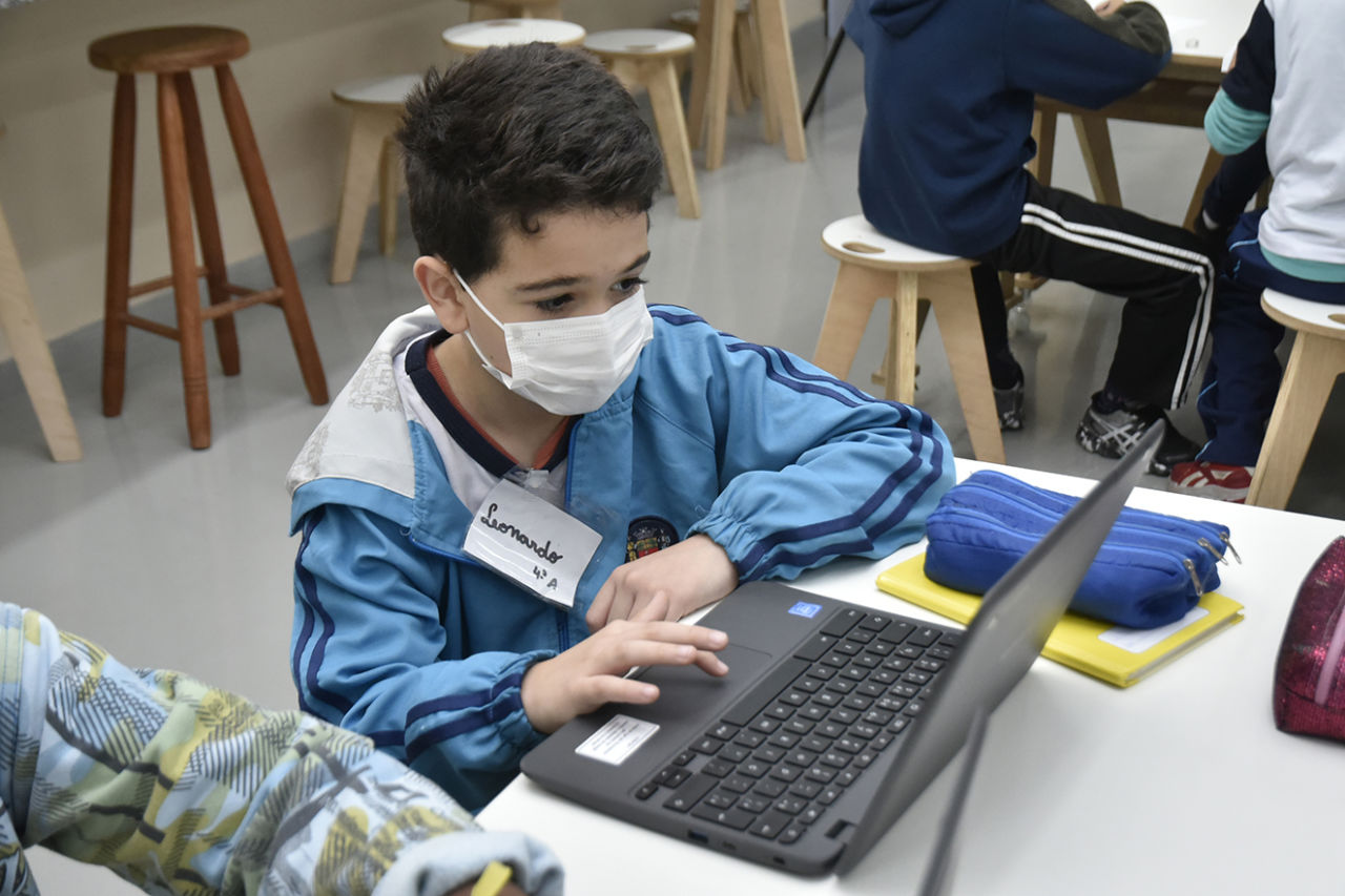 DESCRIÇÃO DA IMAGEM
Aluno veste uniforme azul, usa máscara de proteção facial branca. Ele está sentando em frente a uma mesa branca e mexe em um notebook. Há materiais como estojo e caderno na mesa também.