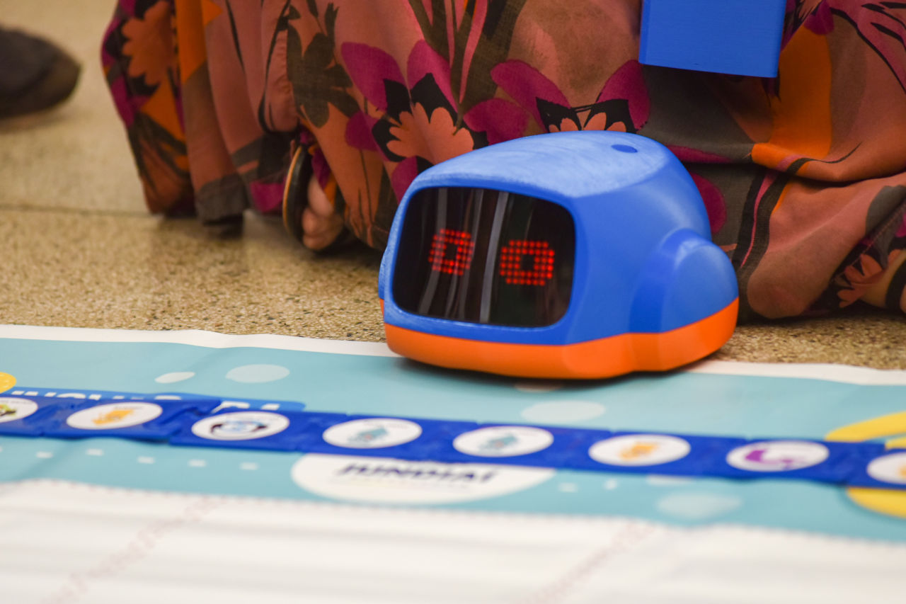 DESCRIÇÃO DA IMAGEM
No chão, há um tapete branco e azul claro, com um robô azul, com base laranja, com olhos, ele está apoiado no chão. Também há peças azuis com adesivos em uma fileira.