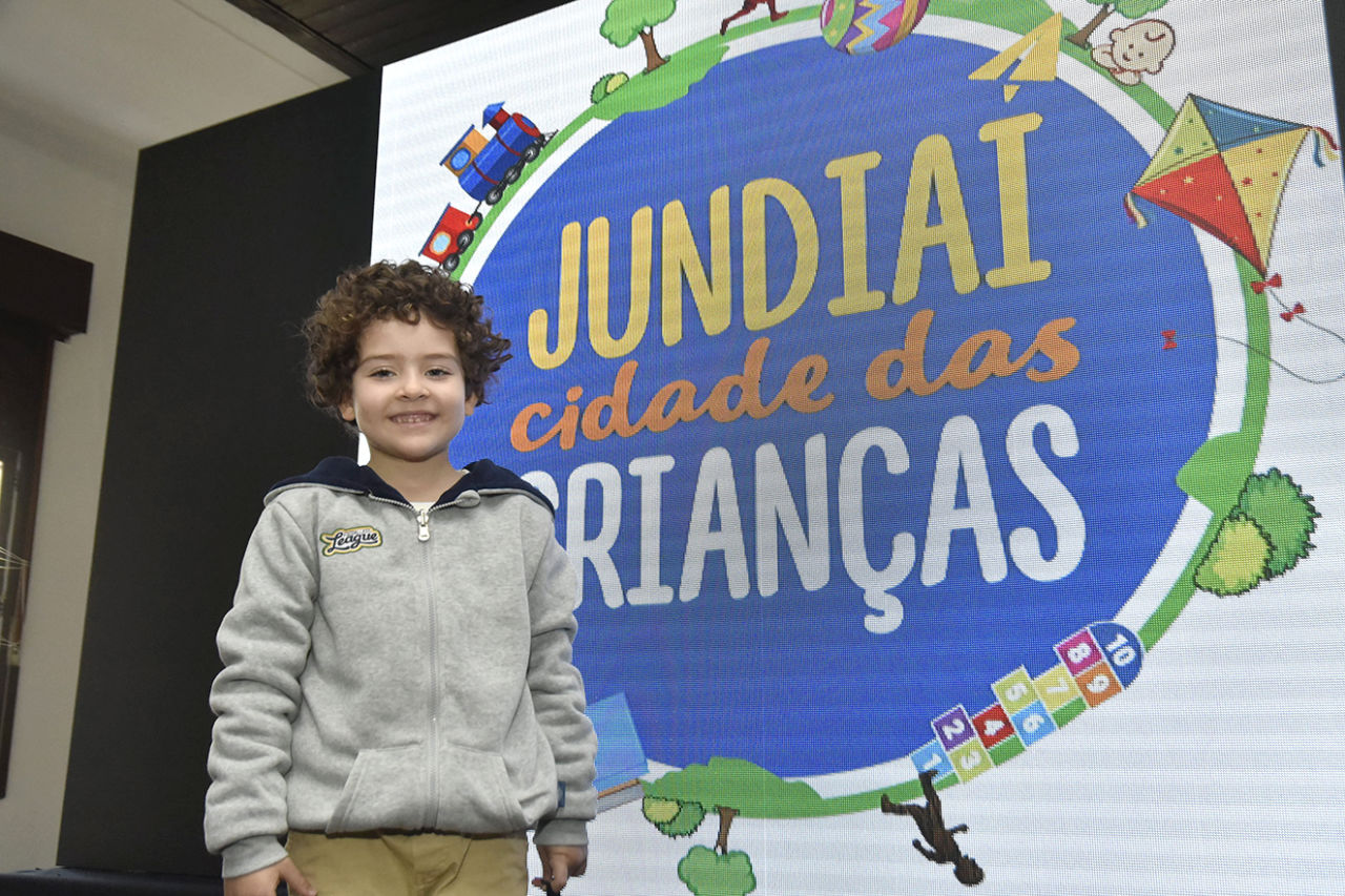 Rafael sorri para a foto. Ele está em frente a um painel com os dizeres: Jundiaí Cidade das Crianças 