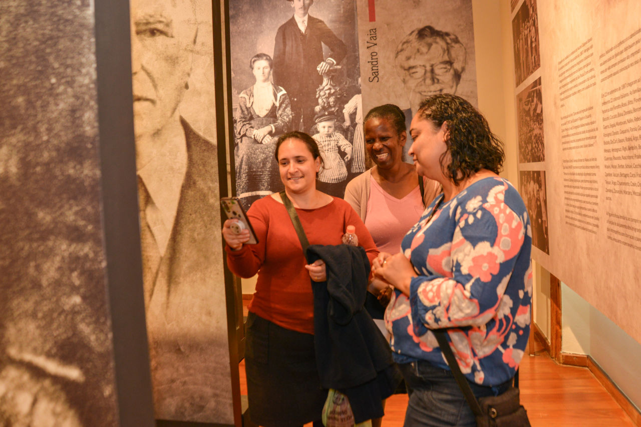 Três mulheres usando blusas de frio interagem, sorridentes, em sala da Pinacoteca Municipal, uma delas segurando celular para o qual todas olham, em meio a painéis de fotos em preto e branco