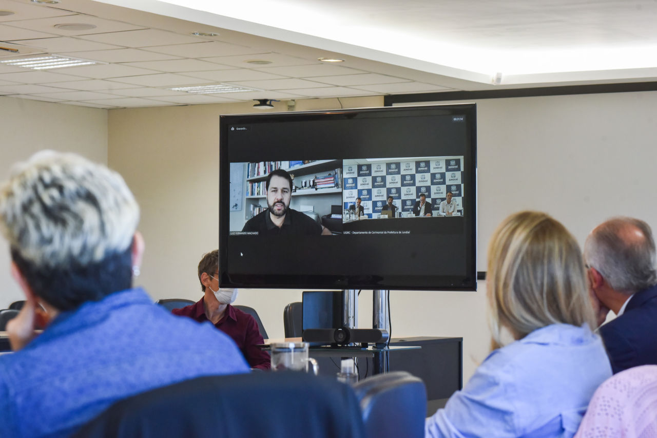 DESCRIÇÃO DE IMAGEM:  Em tela de TV, prefeito Luiz Fernando Machado realiza videoconferência com gestores, na sala de situação da Prefeitura de Jundiaí