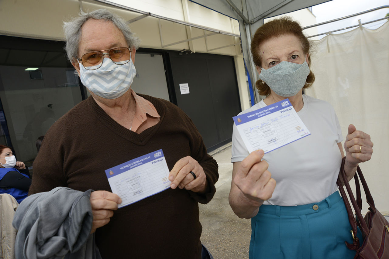 DESCRIÇÃO DE IMAGEM: Casal de idosos apresentam cartões de vacinação com registro da dose adicional após receber a vacina em UBS