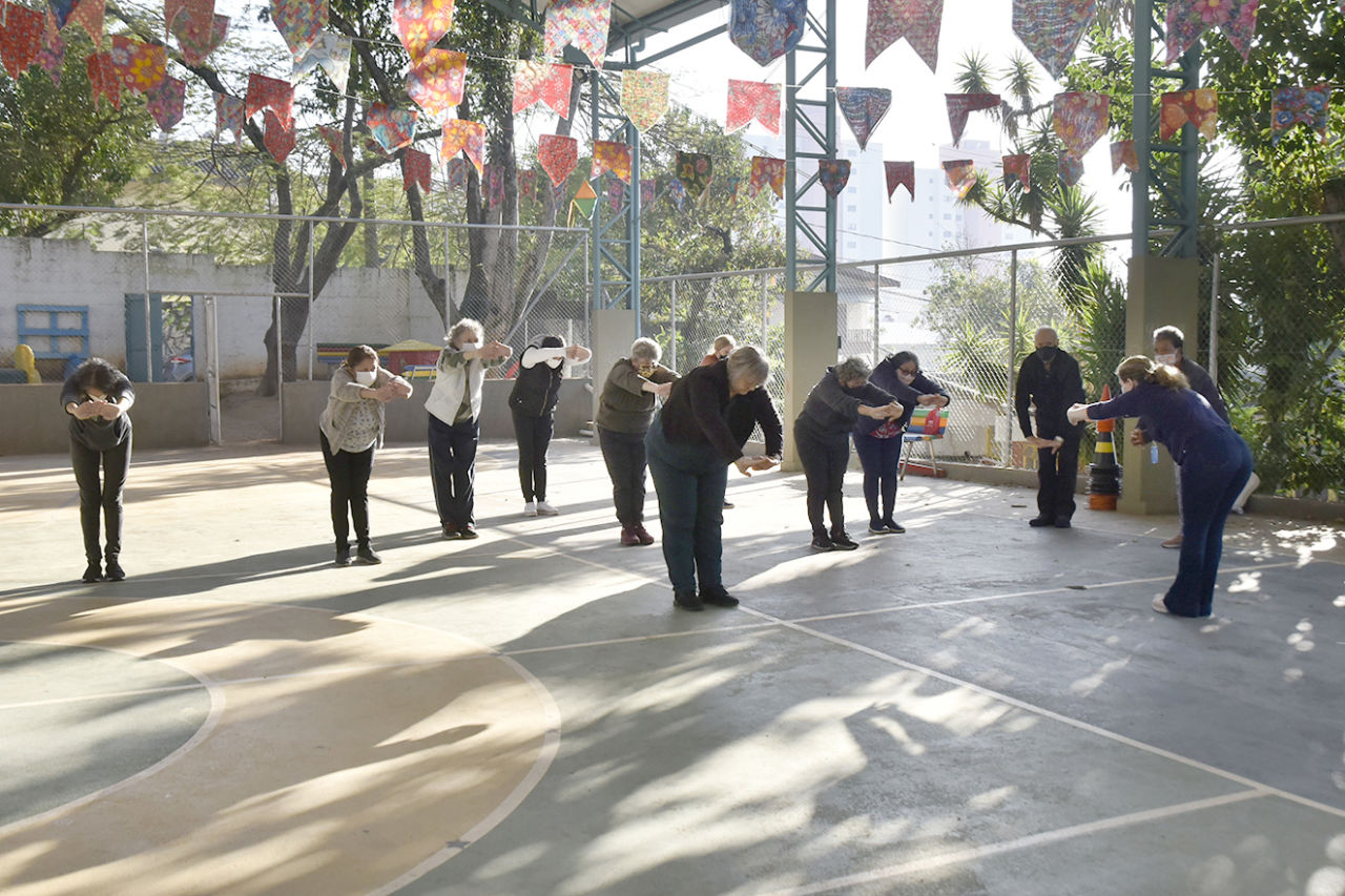 DESCRIÇÃO DA IMAGEM: Em quadra esportiva coberta da EMEB Rotary, grupo de pessoas pratica atividades físicas denominada Lian Gong, com movimentos de respiração e corpo integrados.