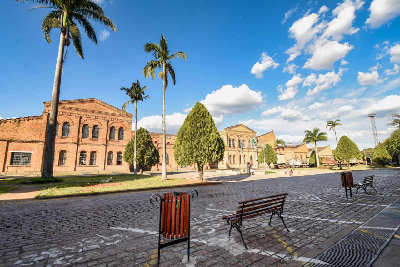Praça central do Complexo Fepasa, onde se veem bancos e lixeiras de madeira, e prédios históricos ao fundo com tijolos aparentes, pinheiros, palmeiras e céu com nuvens