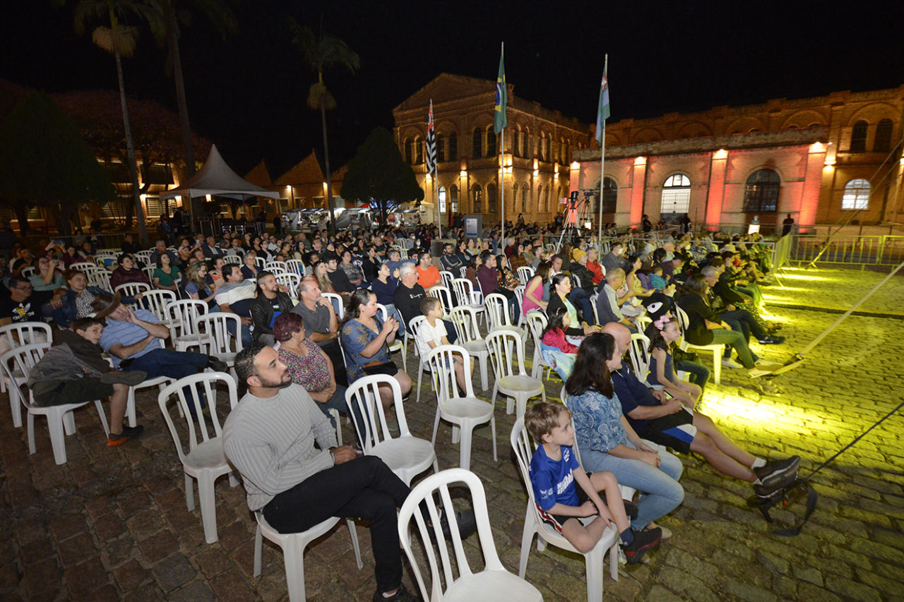 Foto noturna da praça central do Complexo Fepasa com pessoas sentadas em cadeiras de plástico brancas, com prédios históricos de tijolos á vista no fundo