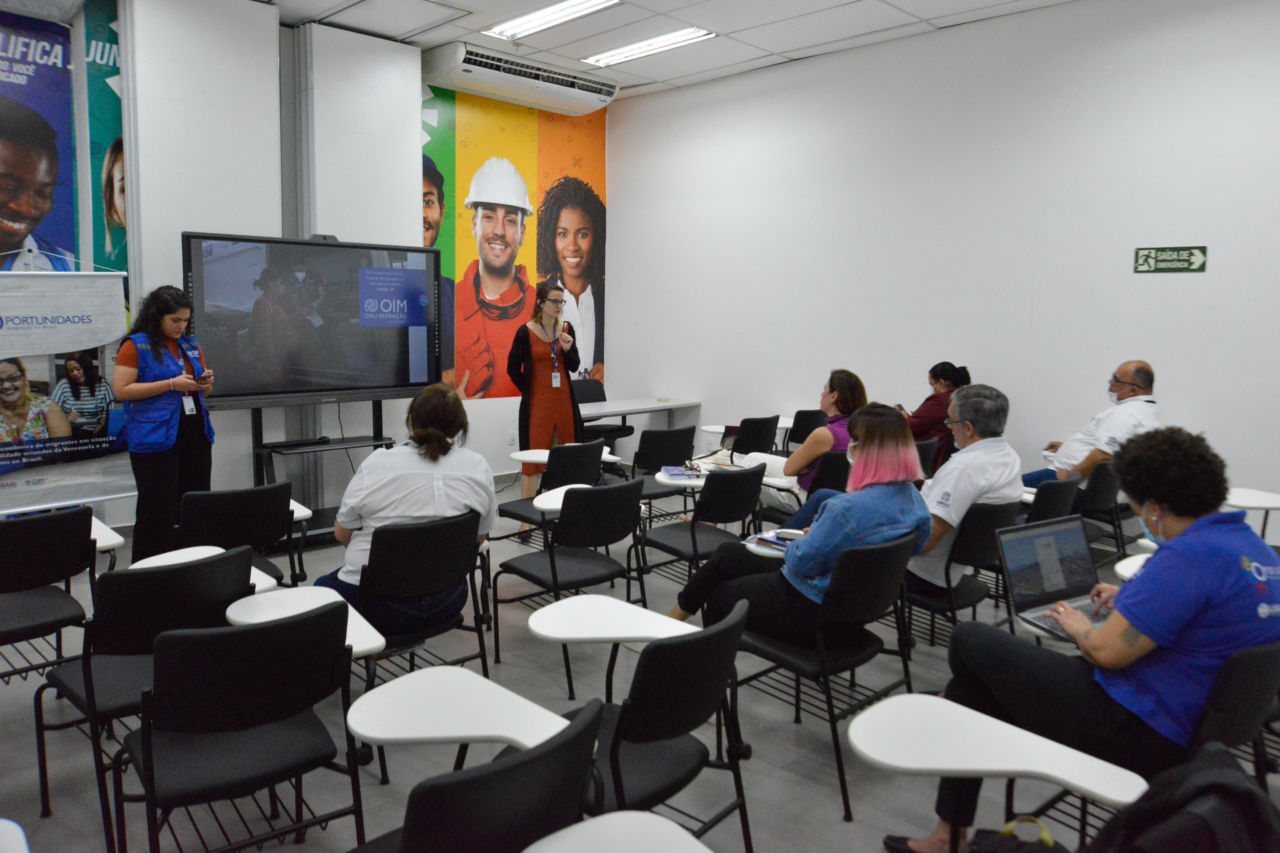 Em uma sala, estão duas mulheres em pé, uma de cada lado de uma tela interativa, com slides sobre a população migrante. Na frente delas tem carteiras e duas pessoas sentadas olhando para elas.