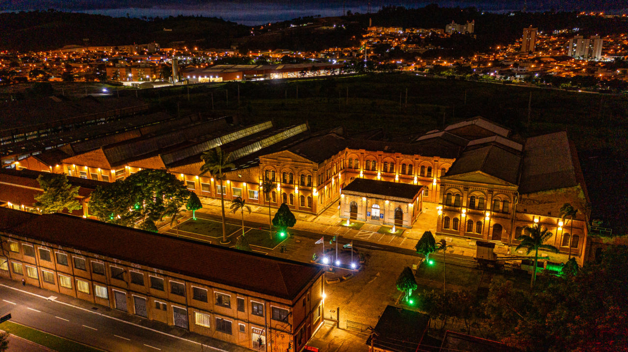 Foto aérea e noturna do Complexo Fepasa iluminado, com outros prédios ao fundo, e iluminação cênica verde em torno de árvores e outras vegetações em vão central