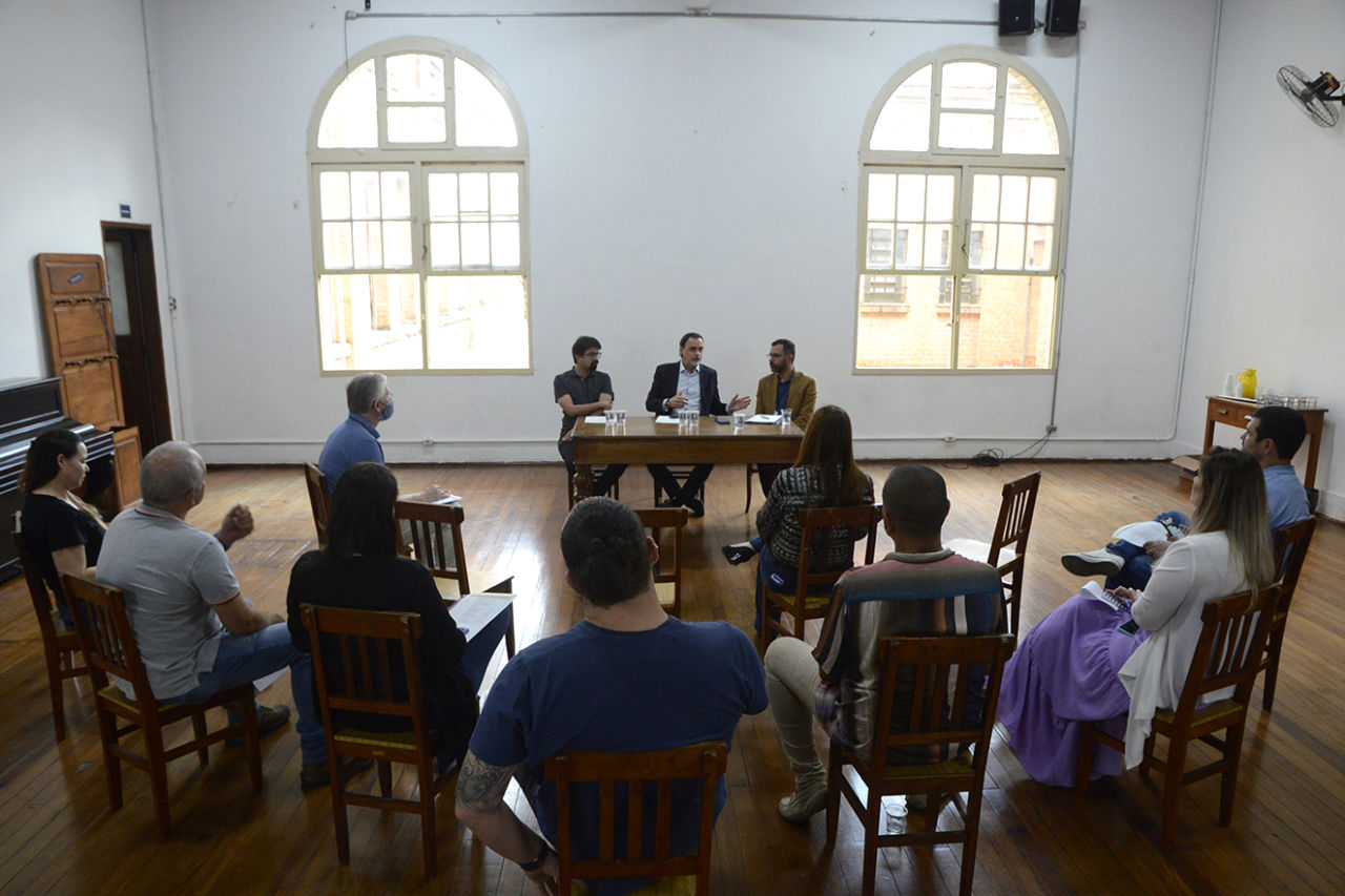 Sala de reunião, com pessoas diversas sentadas em cadeiras de madeira, voltadas para mesa onde se encontram posicionados três homens dialogando, com duas janelas ao fundo