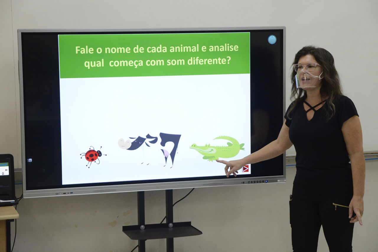 DESCRIÇÃO DA IMAGEM
Professora mexe em tela interativa, que apresenta desenhos de animais e pode-se ler: Fale o nome de cada animal e analise qual começa com som diferente?