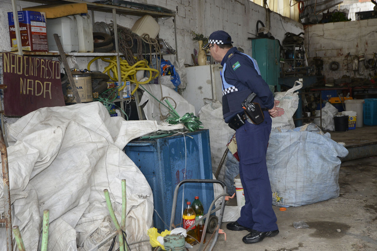 DESCRIÇÃO DA IMAGEM: Guarda municipal com uniforme azul observa material em local de armazenagem de material reciclado