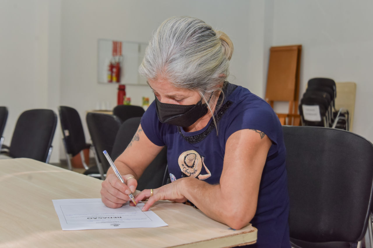 Mulher de cabelos grisalhos e presos, usando máscara facial preta, escrevendo com caneta esferográfica em folha de papel, sentada em cadeira preta e escrevendo em mesa de madeira