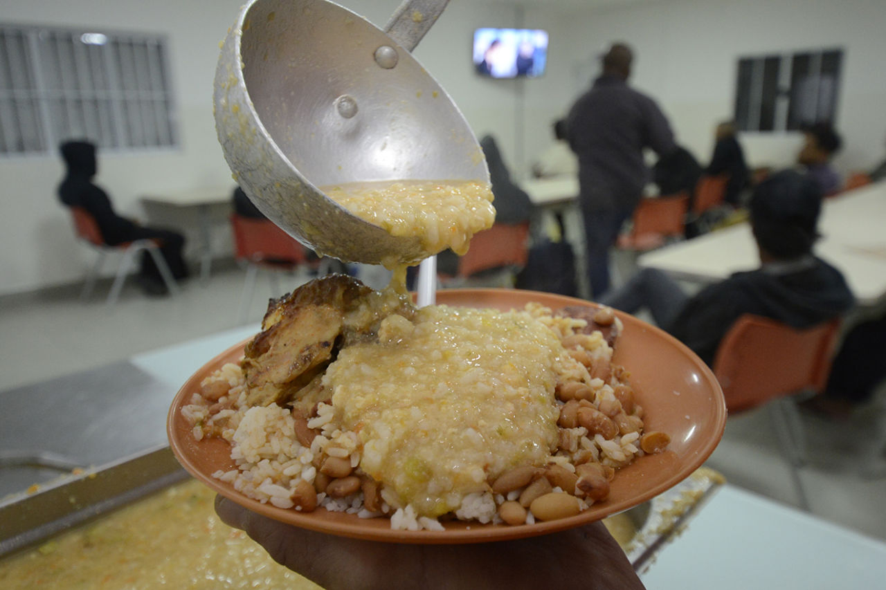 Detalhe de pessoa colocando creme de milho sobre prato com arroz, feijão e carne, com pessoas sentadas em refeitório ao fundo