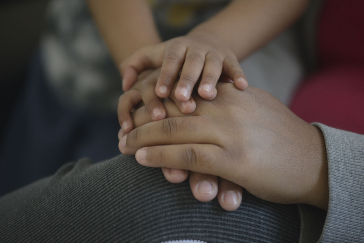 Detalhe de mãos de duas pessoas, uma adulta e uma criança, apoiadas umas sobre as outras, sobre joelho dobrado de uma delas