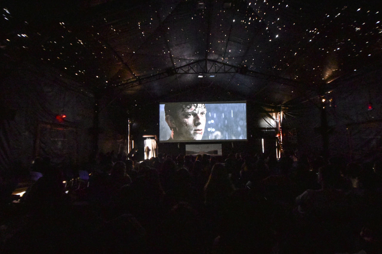 Sala de cinema, com silhuetas de pessoas sentadas na plateia, e tela iluminada em que aparece o ator Tom Holland