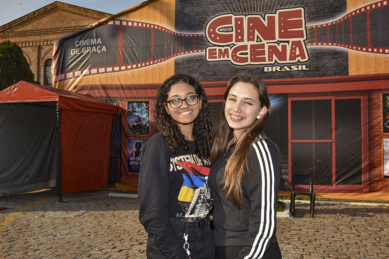 Duas jovens em foto posada e sorrindo, em frente a uma tenda, em que está escrito "Cine em Cena Brasil - Cinema de graça"