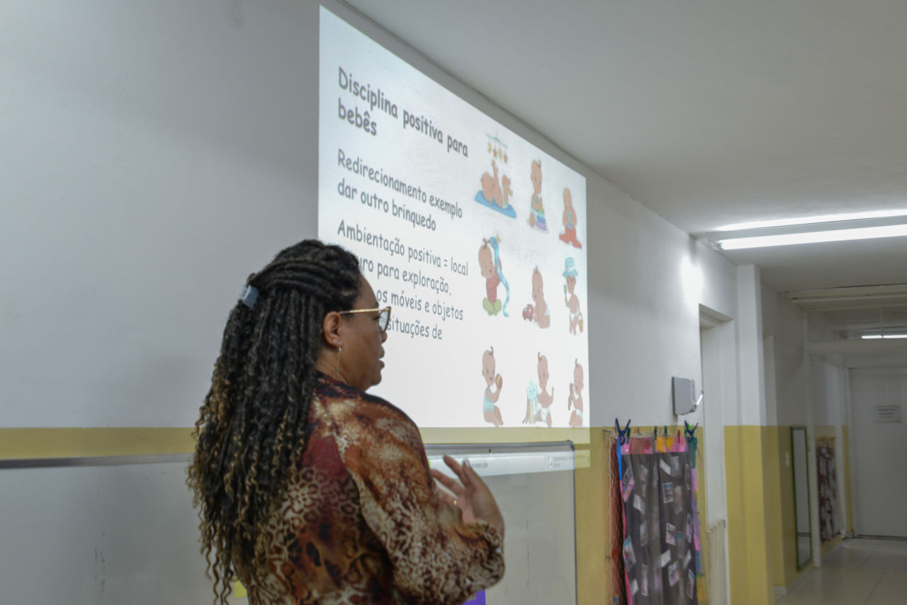A assistente social Isabete Ferreira, está de lado para a foto, olhando para os pais. Na parede ao seu lado, passam os slides sobre "Educação Não Violenta" em apresentação para os pais. Texto no slide: Disciplina positiva para bebês.