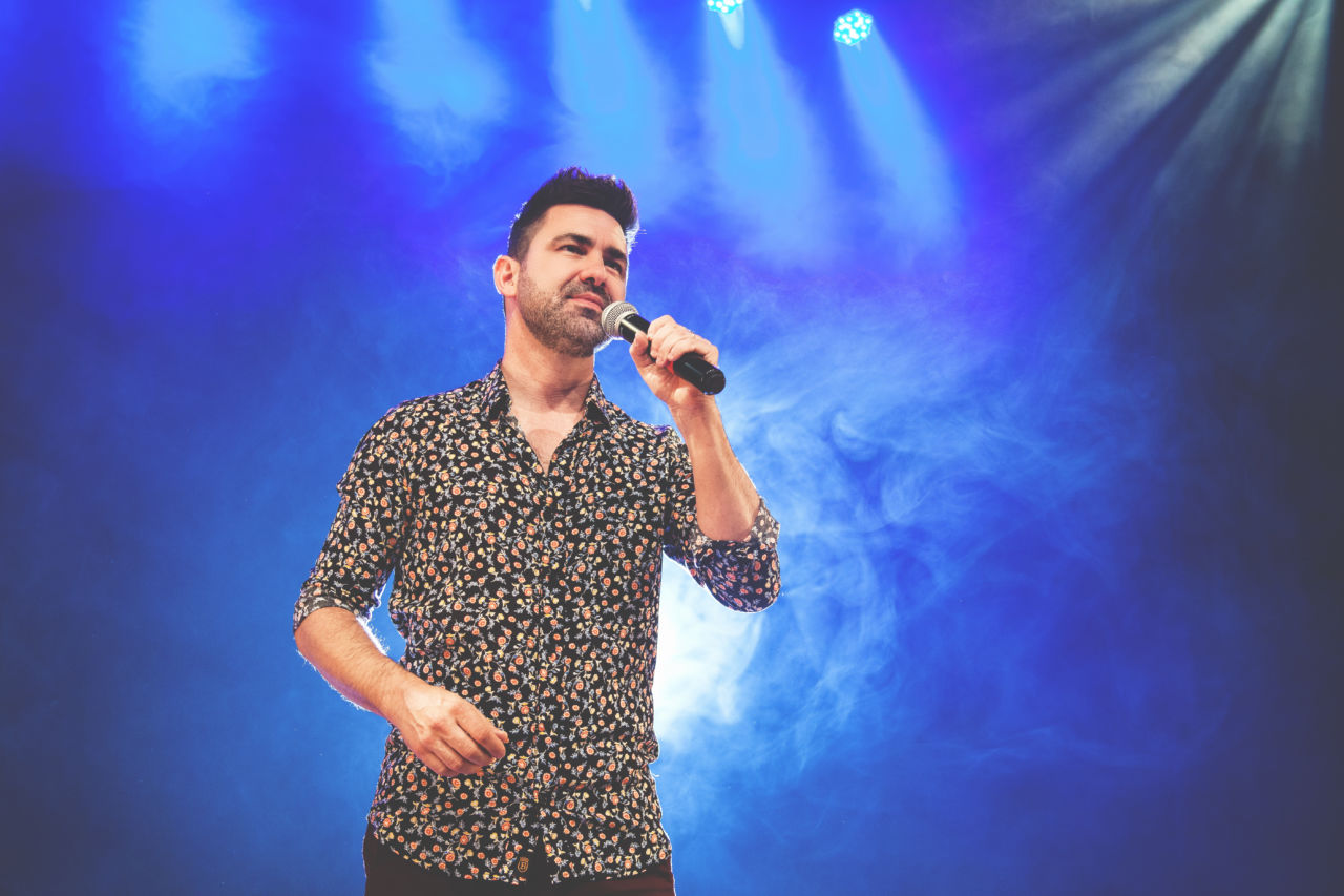 Homem de cabelo curto, barba e camisa florida canta em em palco com iluminação e fumaça, segurando microfone sem fio