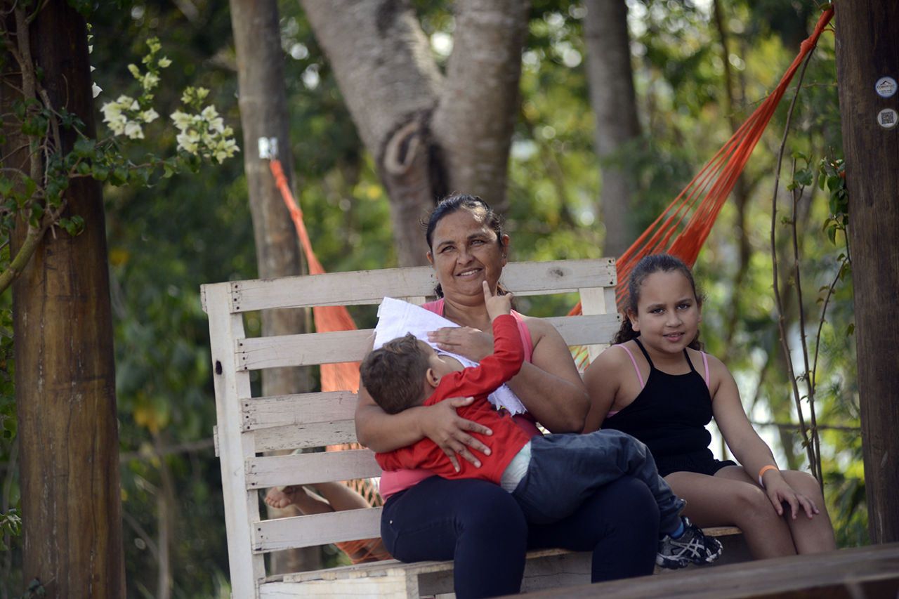 Mulher amamentando um filho, com uma filha ao lado, sentados em banco de madeira, sob a sombra de árvores e com rede ao fundo