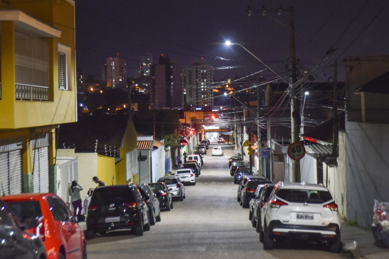 DESCRIÇÃO DA IMAGEM: foto de rua Clélia, na Ponte São João, iluminada com lâmpadas em LED. Luz clara ilumina veículos estacionados nos dois lados da via
