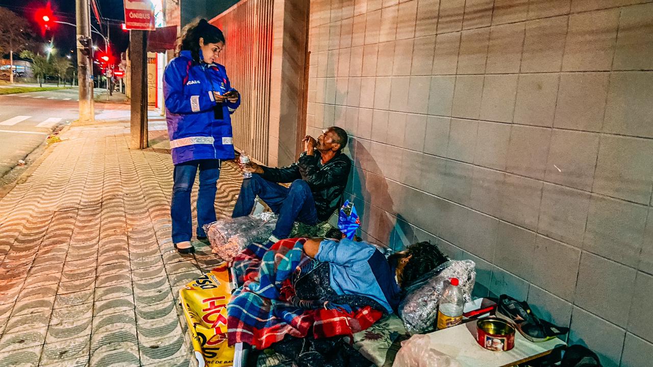 Mulher com casaco de uniforme, em pé,  conversa com dois homens, um sentado e outro deitado, sobre a calçada, junto a cobertores, latas, garrafas e outros pertences