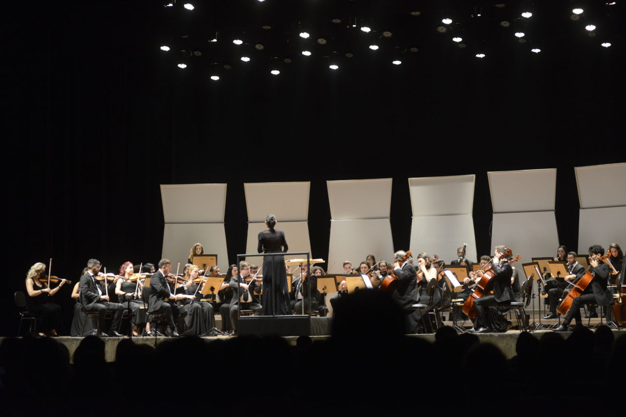 Palco do teatro Polytheama com músicos e instrumentos de orquestra, e mulher regente ao centro, em pé e de costas, com detalhes da iluminação no teto, placas brancas decorativas, e silhuetas escuras do público sentado na plateia