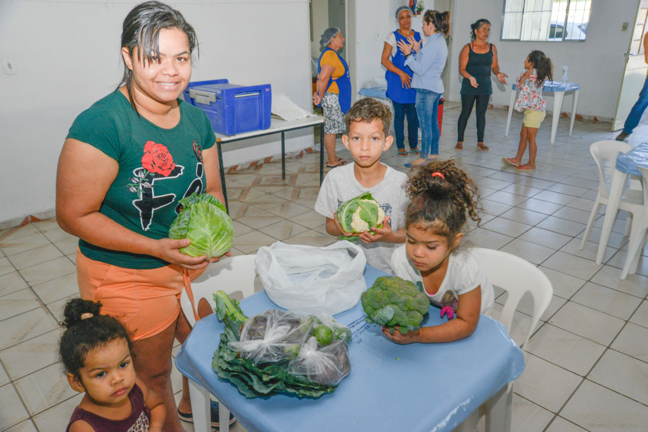 Mulher e três crianças no entorno de uma mesa de plástico, onde apoiam verduras e legumes que tiraram de uma sacola plástica